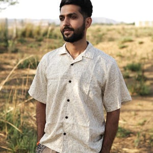 Peut inclure: Chemise à manches courtes crème avec un motif de feuilles répété et des boutons noirs. La chemise a un col classique. La personne porte un short beige avec un motif en spirale sur la poche. Le tissu est léger.