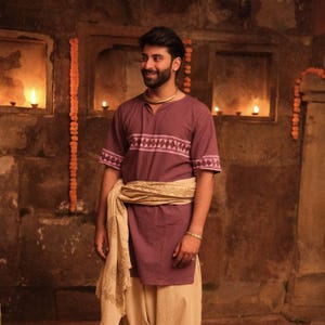 May include: A person wearing a maroon tunic with a geometric design across the chest and sleeves, paired with beige pants. A beige scarf is draped around the waist. The person is wearing a gold necklace and bracelet.