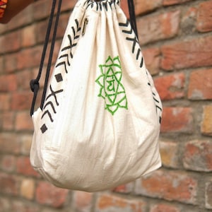 May include: Off-white drawstring backpack with black geometric patterns and a green embroidered design. The bag has a zippered pocket and black drawstrings. The bag is made of a soft, textured fabric.