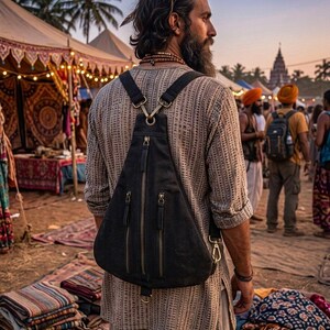 Puede incluir: Una mochila de lona negra con tres compartimentos con cremallera y herrajes de color latón. La bolsa de forma triangular se lleva sobre el hombro. La imagen está tomada en un mercado al aire libre con tiendas de campaña y gente al fondo.
