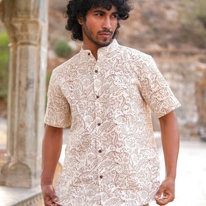 Puede incluir: Una camisa crema de manga corta y pantalones a juego con un estampado de cachemira marrón. La camisa tiene cuello mandarín y cierre de botones. El atuendo lo lleva una persona con cabello rizado oscuro.