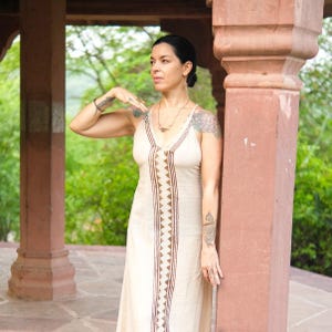 May include: A sleeveless, floor-length cream-coloured dress featuring a vertical brown geometric pattern running down the centre. The garment has a V-neckline with thin straps and a fitted bodice that flows into a straight skirt silhouette.