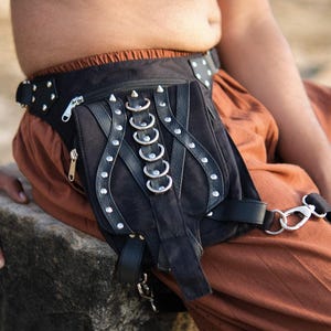 Könnte beinhalten: Schwarze Hüfttasche mit silbernen Nieten, Reißverschlüssen und Metallringen. Die Tasche hat eine Klappe und verstellbare Riemen. Die Tasche wird um die Taille getragen und besteht aus einem Wildleder ähnlichen Material.