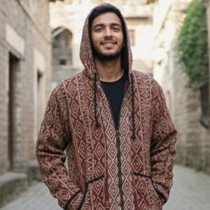 Pode incluir: Casaco com capuz com um padrão geométrico em tons de castanho e bege. O casaco tem fecho de correr completo, capuz e bolsos. A pessoa na imagem está a usar uma camisa preta por baixo.