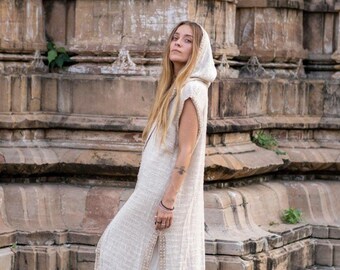 May include: A sleeveless, hooded, floor-length cream-colored robe with a textured weave. The robe has a high slit on the side and is trimmed with a lighter beige border. The person is standing in front of a stone building.