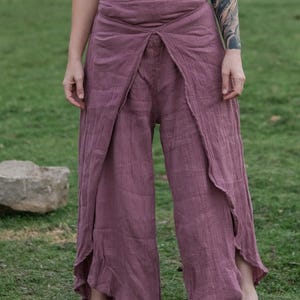 Pode incluir: Calças largas de cor rosa seco com um design de sobreposição frontal. A peça é feita de um tecido texturizado com trama visível, apresenta uma cintura alta e pernas soltas e fluidas que se estendem até aos tornozelos com aberturas.