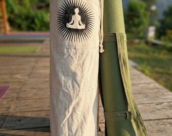 Pode incluir: Uma bolsa de tapete de yoga em lona natural com um design de explosão solar preta e uma figura sentada. Um tapete de yoga verde com uma alça de transporte está ao lado da bolsa. A bolsa tem zíper e cordão.
