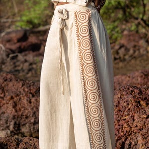 May include: Cream-colored linen skirt with a brown spiral design along the side. The skirt has a tie waist and a matching brown border at the hem. The top is a cream-colored crop top with a geometric pattern.