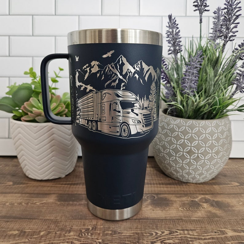 May include: A navy blue stainless steel travel mug with a laser engraved design of a semi-truck driving through a mountain landscape.