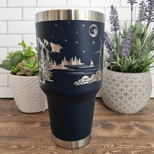 May include: A navy blue stainless steel tumbler with a laser-etched design of a lake scene with mountains, trees, a sailboat, a moon, and stars. The tumbler has a silver lid.