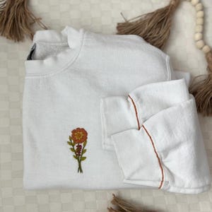 May include: White sweatshirt with a small embroidered orange flower on the front. The sweatshirt has a brown trim around the sleeves.