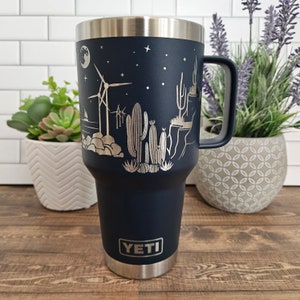 May include: A navy blue YETI tumbler with a silver design of cacti, windmills, and a crescent moon. The tumbler has a stainless steel lid and handle.