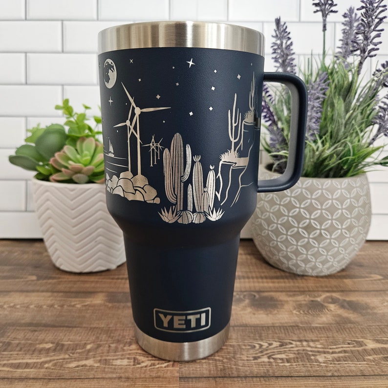 May include: A navy blue YETI tumbler with a silver design of cacti, windmills, and a crescent moon. The tumbler has a stainless steel lid and handle.