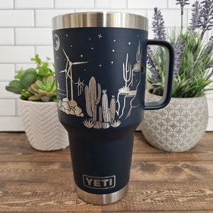 May include: A navy blue YETI tumbler with a stainless steel lid and handle. The tumbler features a laser-etched design of a desert landscape with cacti, windmills, and a crescent moon.