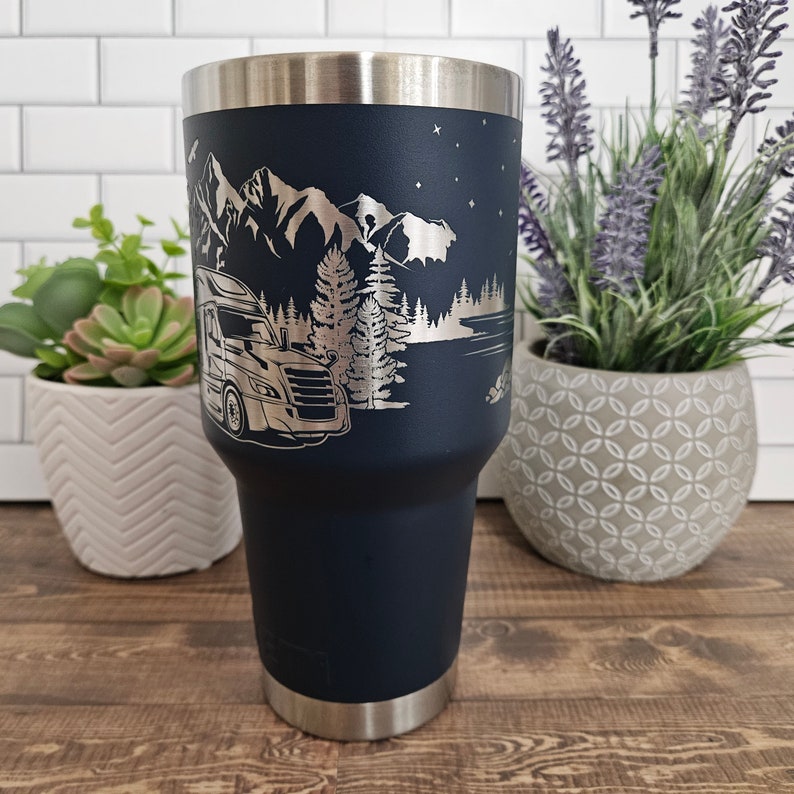 May include: A navy blue stainless steel tumbler with a laser-etched design of a semi-truck driving through a mountain landscape with trees and a lake. The design is in silver.
