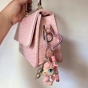 Peut inclure: Un sac à main rose clair avec une texture gaufrée façon crocodile et une poignée supérieure. Un mousqueton en forme de cœur est attaché sur le côté, avec un porte-clés perlé représentant une petite figurine de caniche rose aux grands yeux bleus.