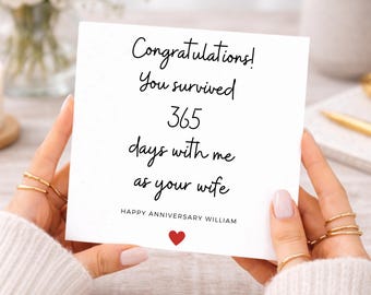Op de afbeelding: Een vierkante witte wenskaart met zwarte tekst die luidt: Congratulations! You survived 365 days with me as your wife. Daaronder staat HAPPY ANNIVERSARY WILLIAM, gevolgd door een klein rood hartpictogram in het midden onderaan de kaart.