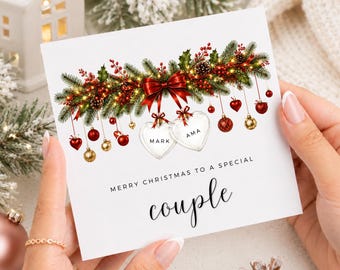 May include: A square white greeting card featuring a festive illustration of evergreen branches, red berries, pinecones, and a red bow. Hanging from the branches are red and gold ornaments and two heart-shaped charms labelled MARK and AMA. Text reads: MERRY CHRISTMAS TO A SPECIAL couple.