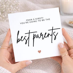 Op de afbeelding: Een vierkante witte wenskaart met de zwarte tekst FRAN & KIRSTY YOU'RE GOING TO BE THE best parents in een combinatie van schreefloze en scriptlettertypes. Een klein, effen roségouden hartje is onder de tekst op de kaart gecentreerd.