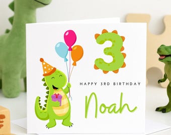 May include: A green dinosaur holding a purple gift box with a party hat on its head. The dinosaur is surrounded by colourful balloons. The text "Happy 3rd Birthday Noah" is written on the card.