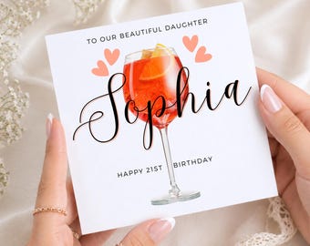 May include: A white birthday card with the text "TO OUR BEAUTIFUL DAUGHTER" and "HAPPY 21ST BIRTHDAY". The card features a watercolour illustration of a cocktail and the name "Sophia" in elegant script. Pink hearts are also included.