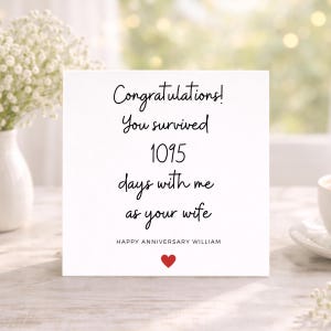 May include: A square white greeting card featuring black text which reads: Congratulations! You survived 1095 days with me as your wife. Below this, it says HAPPY ANNIVERSARY WILLIAM in all caps, followed by a small red heart icon at the bottom centre.