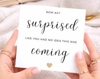 Op de afbeelding: Een vierkante witte kaart met zwarte tekst die NOW ACT surprised LIKE YOU HAD NO IDEA THIS WAS coming leest en een klein gouden hartsymbool onderaan. De kaart wordt vastgehouden door twee handen met meerdere gouden ringen tegen een witte achtergrond.
