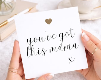 May include: A square white greeting card featuring the black text you've got this mama with a small gold heart icon above the text and a small black x below the text. The card is held in a centred position against a bright, soft background.