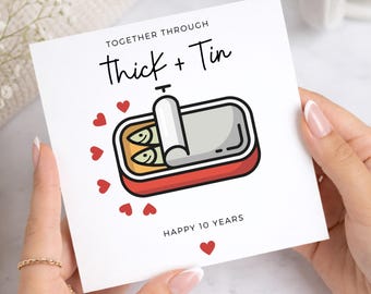 May include: A square greeting card with a white background featuring a cartoon illustration of an open red tin of sardines. The text reads TOGETHER THROUGH Thick + Tin and HAPPY 10 YEARS. Several small red hearts surround the sardine tin.