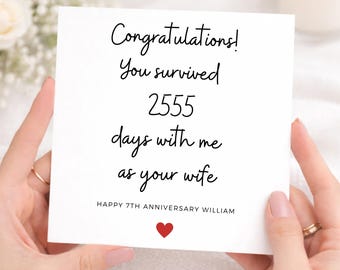 Könnte beinhalten: Eine quadratische weiße Grußkarte mit schwarzem kursivem Text, der lautet: Congratulations! You survived 2555 days with me as your wife. Darunter steht in Großbuchstaben HAPPY 7TH ANNIVERSARY WILLIAM, gefolgt von einem kleinen roten Herzsymbol.