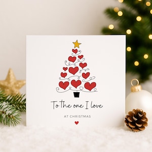 Könnte beinhalten: Eine weiße Weihnachtskarte mit einem Baum aus roten Herzen und einem goldenen Stern. Der Text lautet "To the one I love AT CHRISTMAS" mit einem kleinen roten Herz darunter. Ein goldener Stern und ein Tannenzapfen sind im Hintergrund.