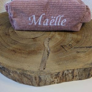 Può includere: Astuccio in velluto a coste rosa antico con cerniera bianca e il nome "Maëlle" ricamato in corsivo bianco. L'astuccio è appoggiato su una superficie di legno rustico.