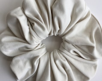 May include: A large, ivory-colored scrunchie made of soft fabric. The scrunchie is circular with a gathered design, creating a voluminous appearance. The fabric has a subtle sheen, suggesting a velvet or similar texture. This hair accessory is suitable for various hairstyles.