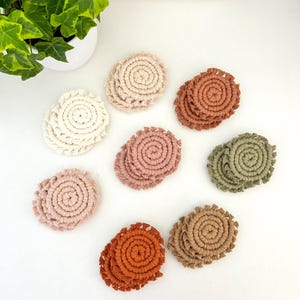 May include: Eight handmade macrame coasters in various colors, including beige, brown, green, and orange. The coasters are circular and have a fringed edge.
