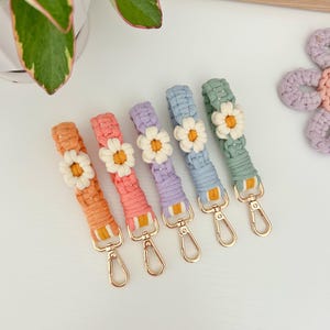 Könnte beinhalten: Fünf bunte gehäkelte Schlüsselanhänger mit Gänseblümchen-Anhängern und goldenen Schlüsselringen. Die Schlüsselanhänger sind in Orange-, Rosa-, Lila-, Blau- und Grüntönen gehalten.