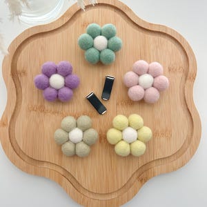 Puede incluir: Cinco pinzas para el pelo de fieltro en forma de flor en varios colores, incluyendo verde, morado, rosa, amarillo y beige. Cada flor tiene un centro blanco y está unida a una pinza de metal plateado.