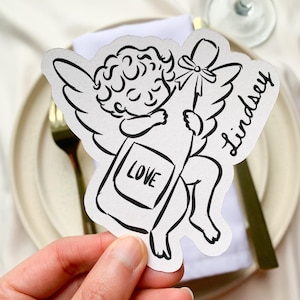 May include: A black and white illustration of a cherub holding a champagne bottle with a bow. The word "LOVE" is on the bottle. The name "Lindsey" is written on the right side of the cherub. The cherub is on a white background.
