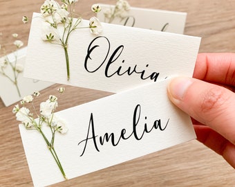 May include: Two white place cards with calligraphy names "Olivia" and "Amelia" decorated with small white flowers.