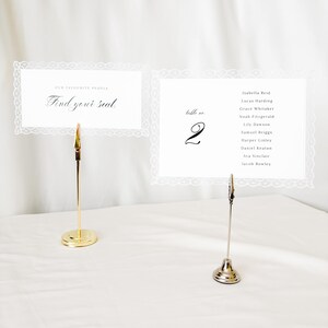 May include: Two rectangular white paper cards with intricate lace-style borders held by tall gold-toned metal stands. One card reads "OUR FAVOURITE PEOPLE Find your seat." The other card displays "table no. 2" and a list of ten names in black text.