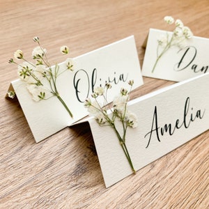 May include: Three white place cards with calligraphy names "Olivia", "Amelia", and "Dan" with small white flowers on each card.