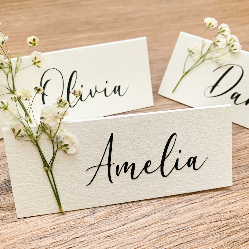 May include: Three white place cards with black calligraphy names, Olivia, Amelia, and Dan, with sprigs of white baby's breath flowers.