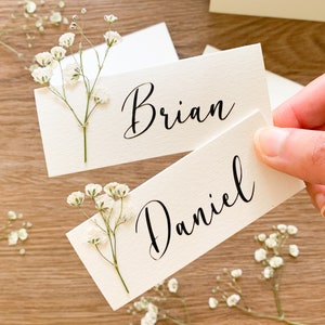 May include: Two white place cards with calligraphy names "Brian" and "Daniel" decorated with small white flowers.
