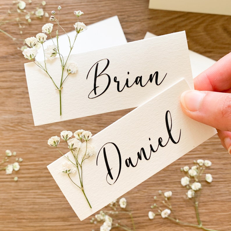 May include: Two white place cards with calligraphy names "Brian" and "Daniel" decorated with small white flowers.