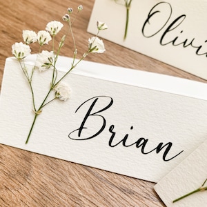 May include: White place card with the name "Brian" written in black cursive script. A sprig of white baby's breath flowers is placed on the left side of the card.