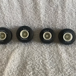 May include: Four black Goodyear Eagle tires with white rims. The tires are arranged in a row, showcasing their tread and the "GOOD YEAR" and "EAGLE" branding in white lettering. The rims have a series of small holes.