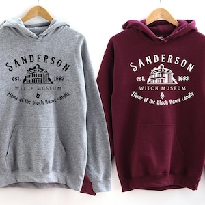 May include: Two hooded sweatshirts with a graphic print of a house and the text "SANDERSON est. 1693 WITCH MUSEUM Home of the black flame candle". The sweatshirt on the left is grey and the sweatshirt on the right is maroon.