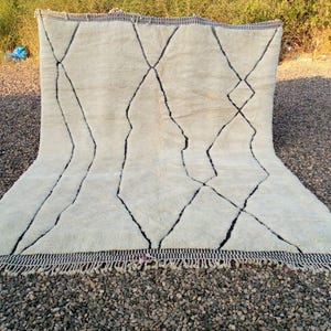 May include: Cream-colored rug with a geometric black diamond pattern. The rug has a thick, textured pile and a fringe along the bottom edge. The design features bold, irregular black lines creating a striking contrast against the neutral background.