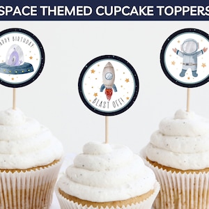 May include: Three space-themed cupcake toppers. The first topper features a purple alien in a flying saucer with the text "Happy Birthday". The second topper features a rocket with the text "Blast Off!" The third topper features an astronaut with a white helmet and a blue spacesuit.