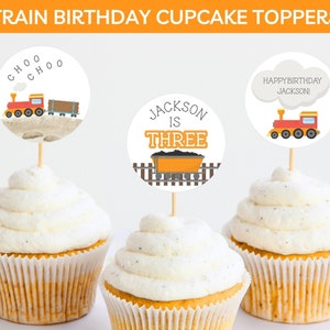 May include: Three cupcake toppers featuring a train theme. One topper says "Choo Choo", another says "Jackson is Three", and the last one says "Happy Birthday Jackson".