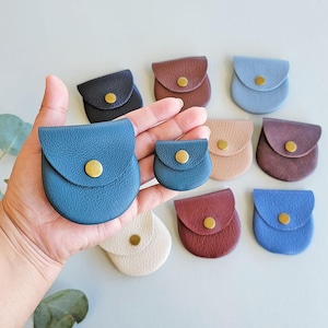 May include: A hand holds two small, blue leather coin purses with gold snap closures. Several other small, round leather coin purses in various colors, including brown, pink, and blue, are arranged in the background.